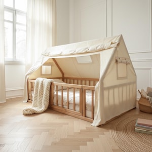 May include: A wooden house-shaped bed frame with a cream-colored canvas canopy. The bed has a wooden railing and a white mattress. A cream-colored knitted blanket is draped over the bed. A woven basket and a stack of books are nearby.