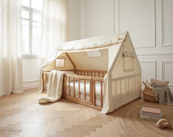 Könnte beinhalten: Ein holzfarbenes Hausbett mit cremefarbenem Segeltuchdach. Das Bett hat ein Holzgeländer und eine weiße Matratze. Eine cremefarbene Strickdecke ist über das Bett drapiert. Ein geflochtener Korb und ein Stapel Bücher befinden sich in der Nähe. Das Bett ist ca. 160 cm lang.