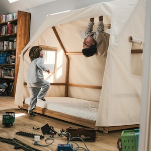 Könnte beinhalten: Zwei Kinder spielen in einem weißen Zelt aus Segeltuch mit einem Holzrahmen. Ein Kind steht auf dem Bett, während das andere kopfüber von der Oberseite des Zeltes hängt. Das Zelt ist in einem Raum mit Holzboden und einem Bücherregal im Hintergrund aufgebaut.