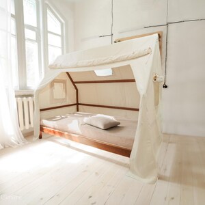 Op de afbeelding: Een wit hemelbedframe met een houten basis en een witte stoffen kap. Het bed is opgemaakt met een wit laken en een beige kussen.