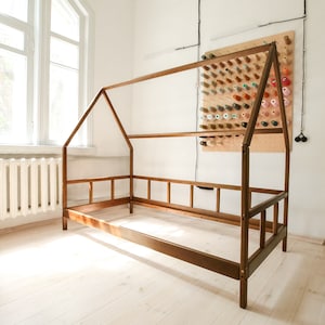 May include: A wooden bed frame in the shape of a house with a white wall and a pegboard with colorful spools of thread in the background.