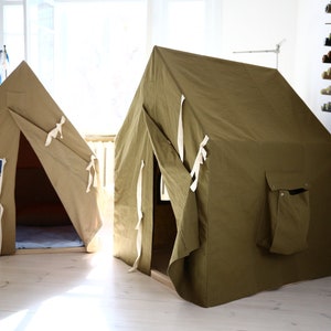 May include: Two canvas play tents. One is tan with a blue and white logo that says "Mountaineer". The other is olive green with a side pocket. Both have white tie closures and are set on a light wood floor.
