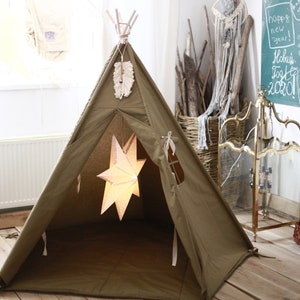 Op de afbeelding: Een groene canvas tipi tent voor kinderen met een wit stervormig licht dat erbinnen hangt. De tipi is opgezet in een kamer met houten vloer en een witte muur met een schoolbord.