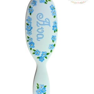 May include: A light blue baby brush with the name "Ava" in light blue script. The brush is decorated with hand-painted blue forget-me-not flowers and green leaves. The brush has a handle and is oval-shaped.