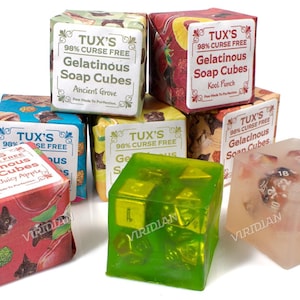 May include: Six colorful boxes of TUX'S 98% Curse Free Gelatinous Soap Cubes. The boxes feature different designs and scents, including Ancient Grove, Kool Punch, and Auntie's Juicy Apple. Three clear soap cubes are also shown, each containing a different set of dice.