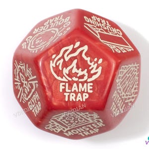 May include: A red, multi-sided die with white text and graphics. The die features the words "FLAME TRAP" and other trap-related terms. The die is designed for tabletop gaming and has a pearlescent finish.