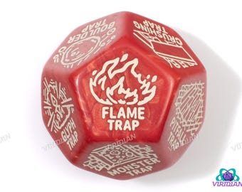 May include: A red, multi-sided die with white text and graphics. The die features the words "FLAME TRAP" and other trap-related terms. The die is designed for tabletop gaming and has a pearlescent finish.