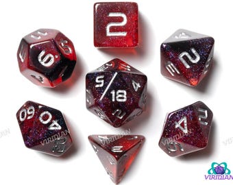 May include: Set of seven polyhedral dice in shades of red and purple with silver numbering. The dice are various shapes, including a cube, a d4, a d6, a d8, a d10, a d12, and a d20. The dice are for tabletop games.