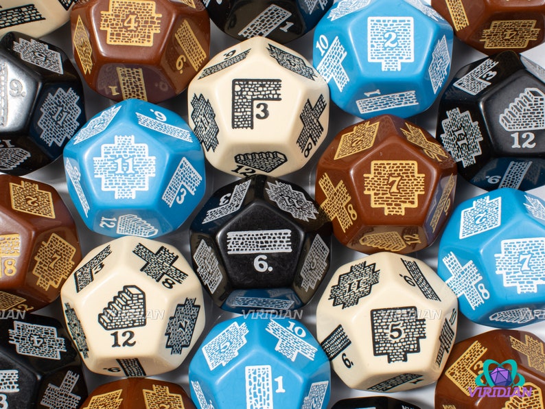 Op de afbeelding: Een verzameling van 12-zijdige dobbelstenen met een kasteelmuurontwerp. De dobbelstenen zijn in verschillende kleuren, waaronder bruin, blauw, zwart en cr&egrave;me. Elke dobbelsteen heeft een ander nummer van 1 tot 12 erop gedrukt.
