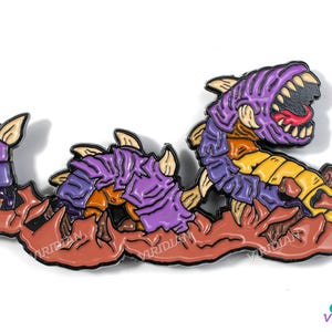 May include: A colorful enamel pin depicting a stylized worm-like creature with a large open mouth. The pin features purple, yellow, orange, and brown colors. The creature is set against a brown background.
