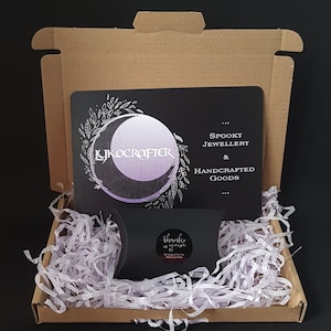 May include: A cardboard box with a black card inside. The card has a moon and floral design with the text "Lykocrafter" and "Spooky Jewellery & Handcrafted Goods". The box is filled with white packing paper and a small black bag with the text "Thank you" on it.