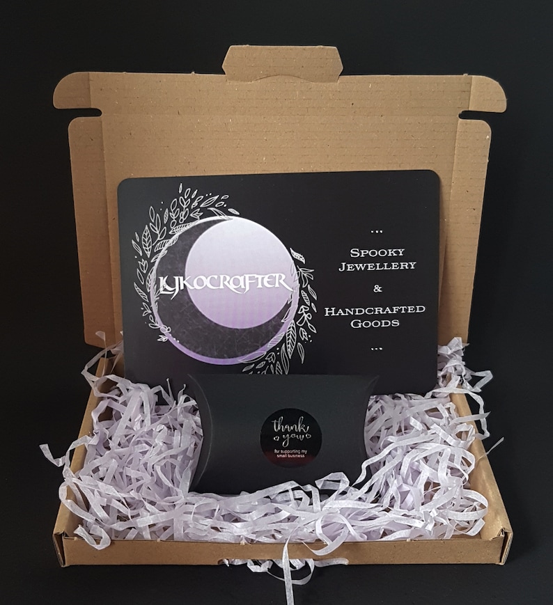 May include: A cardboard box with a black card inside. The card has a moon and floral design with the text "Lykocrafter" and "Spooky Jewellery & Handcrafted Goods". The box is filled with white packing paper and a small black bag with the text "Thank you" on it.
