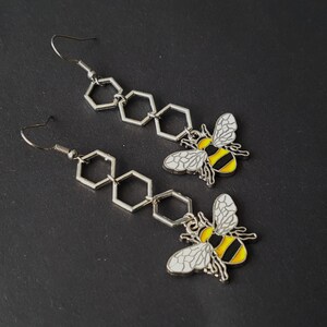 Könnte beinhalten: Ein Paar silberne Ohrringe mit einem Wabenmuster und einem Bienenanhänger. Der Bienenanhänger ist gelb und schwarz mit weißen Flügeln.