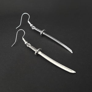 May include: A pair of silver earrings shaped like swords. The swords have a detailed design and are hanging from silver hooks.