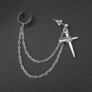 May include: A silver ear cuff with a chain attached to a silver earring with a nail design. The earring has a small silver bead at the top.