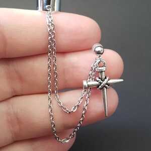 May include: Silver ear cuff with a dangling chain and a silver cross charm with a nail design. The cross is attached to the chain with a small silver bead.