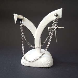 May include: A pair of silver earrings with chains. One earring is a cuff with a chain attached. The other earring is a nail with a chain attached. The earrings are displayed on a white stand.