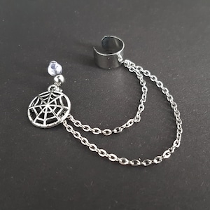 May include: Silver-toned ear cuff with a dangling spiderweb charm and two delicate chains. The ear cuff wraps around the ear, connected to the charm by the chains. A small, clear gem is above the spiderweb charm. The jewelry is set against a dark background.