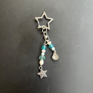 May include: A silver star-shaped keychain with a silver charm of a seashell and a silver star charm. The keychain is decorated with blue and white beads.