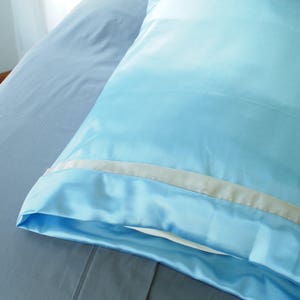 Puede incluir: Una funda de almohada de satén azul claro con ribete blanco. La funda de almohada está sobre una sábana gris.