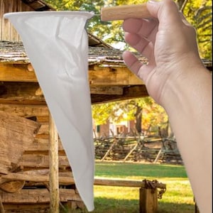 May include: A white, cone-shaped filter with a wooden handle. The filter is held up against a rustic wooden structure, with a blurred background of trees and a building.