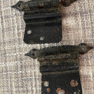 May include: Two black metal door hinges with a worn, aged finish. The hinges have a decorative pointed design at the top and bottom.