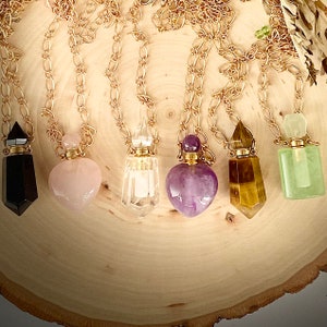 May include: A collection of gold-toned chain necklaces, each featuring a unique gemstone pendant. The pendants include black, pink, clear, purple, brown, and green stones, showcasing various shapes and textures. The necklaces are displayed on a wooden surface.