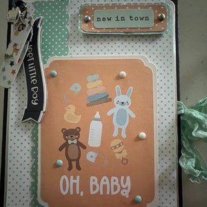 May include: A baby-themed scrapbook with a black cover and a light orange central panel. The panel features illustrations of baby items and the words "OH, BABY". The scrapbook includes the text "new in town" and "meet little boy".