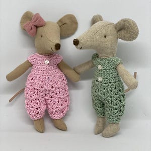May include: Two plush mouse toys wearing crocheted outfits. The mouse on the left is wearing a pink outfit with a pink bow on its head. The mouse on the right is wearing a green outfit.