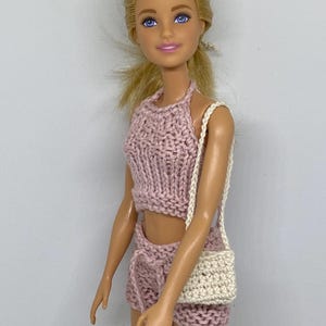 May include: A doll wearing a knitted two-piece outfit in a light pink hue, consisting of a halter top and shorts. The doll also has a small, off-white crocheted purse with a shoulder strap. The doll has blonde hair and blue eyes.
