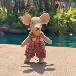 May include: A small, plush mouse wearing a pink crocheted overall with two white buttons. The mouse is standing on a light brown surface with a swimming pool in the background.