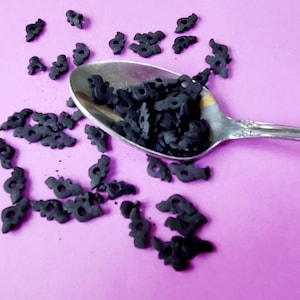 May include: A silver spoon filled with black bat-shaped sprinkles on a pink surface. The sprinkles are scattered around the spoon.