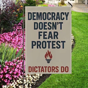 May include: A beige garden flag with the text "DEMOCRACY DOESN'T FEAR PROTEST" in dark blue letters and a flame graphic. Below, in red letters, it reads "DICTATORS DO". The flag is displayed in a garden with pink and white flowers and green grass.