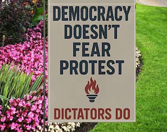 May include: A beige garden flag with the text "DEMOCRACY DOESN'T FEAR PROTEST" in dark blue letters and a flame graphic. Below, in red letters, it reads "DICTATORS DO". The flag is displayed in a garden with pink and white flowers and green grass.
