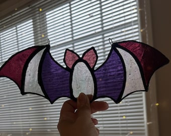 Puede incluir: Un atrapasoles de vidriera con forma de murciélago que presenta segmentos de vidrio en color magenta intenso, blanco y morado, unidos por líneas de soldadura de metal negro. El murciélago tiene dos orejas pequeñas y alas extendidas.