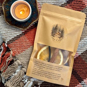 May include: A lit candle in a ceramic holder sits on a plaid blanket next to a kraft paper bag. The bag contains dried orange slices and a cinnamon stick. The bag has a label that says "Cozy Cabin".