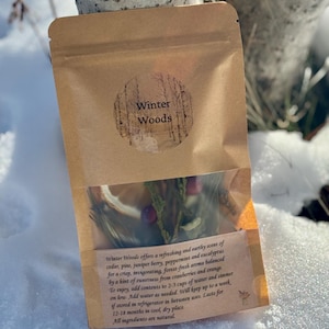 May include: A brown paper bag with a clear window, filled with dried herbs and berries. The bag has a circular label that reads "Winter Woods." Text on the bag describes the contents as cedar, pine, juniper berry, peppermint, and eucalyptus.