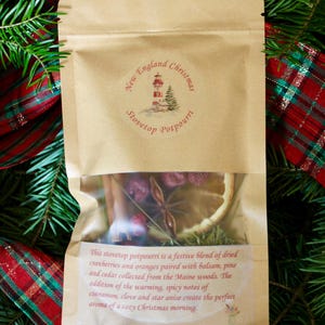 Könnte beinhalten: Ein Kraftpapierbeutel mit Stovetop-Potpourri und einem transparenten Fenster, mit getrockneten Cranberries, Orangenscheiben und Sternanis. Der Beutel ist mit "New England Christmas Stovetop Potpourri" beschriftet und von Tannenzweigen und kariertem Band umgeben.