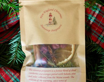 May include: A kraft paper bag of stovetop potpourri with a clear window, featuring dried cranberries, orange slices, and star anise. The bag is labelled "New England Christmas Stovetop Potpourri" and is surrounded by evergreen branches and tartan ribbon.