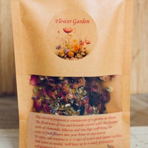 May include: A brown paper bag with a label that says "Flower Garden". The bag contains a mixture of dried flowers and herbs, including rose petals, lavender, chamomile, and hibiscus. The text on the label describes the product as a stovetop potpourri that is reminiscent of a garden in bloom.