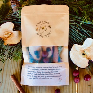 May include: A brown paper bag with a white label that says "A Piece of Maine Stovetop Potpourri". The label has a map of Maine with a red star in the center. The bag contains a mixture of dried cranberries, blueberries, apples, and other ingredients. The text on the label reads: "The stovetop potpourri contains local elements from Maine. Blueberries, cranberries and apples have been harvested from Maine farms and orchards and balsam, cedar and pine sourced from the Maine woods. To enjoy this piece of Maine add contents to 2-3 cups of water and simmer on low."