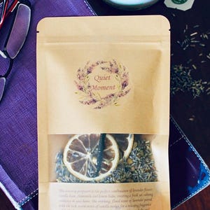 May include: A brown paper bag with a clear window showing dried lavender flowers, lemon slices, and vanilla beans. The bag has a label that says "Quiet Moment" with a watercolor design of lavender flowers. The text on the bag reads: "This sleep sachet is the perfect combination of lavender flower, vanilla bean, chamomile and lemon balm, creating a fresh yet calming ambiance in your home. The soothing, floral scent of lavender paired with the tick warm notes of vanilla makes for a relaxing fragrance while the freshness of chamomile and crispness of lemon balm and lemon slices offer brightness. To enjoy, add contents to 2-3 cups of hot water as needed. Will keep up to a week stored in refrigerator in between use. All ingredients are natural."