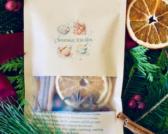 May include: A kraft paper pouch labelled "Christmas Kitchen" with a clear window showing dried orange slices, star anise, and cinnamon sticks. The pouch is surrounded by evergreen branches, dried orange slices, and red berries.