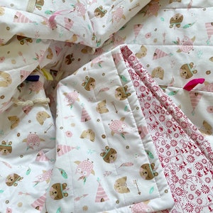 Könnte beinhalten: Eine weiße Babydecke mit einem rosa-braunen Waldtier-Druck. Die Decke ist reversibel und hat auf der anderen Seite einen roten und weißen Blumendruck.