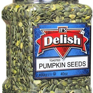 May include: A clear plastic jar filled with toasted pumpkin seeds. The jar has a black lid and a blue and red label that reads "It's Delish" and "Toasted Pumpkin Seeds". The label also includes the text "Pareve" and "40oz".
