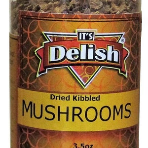 May include: A clear glass jar with a black lid filled with dried, kibbled mushrooms. The label is gold with a red and white logo that says "It's Delish" and the text "Dried Kibbled Mushrooms 3.5oz".