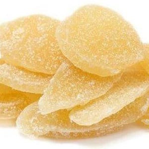May include: A pile of translucent, pale yellow, candied ginger slices. The ginger is glistening and appears to be coated in sugar crystals.