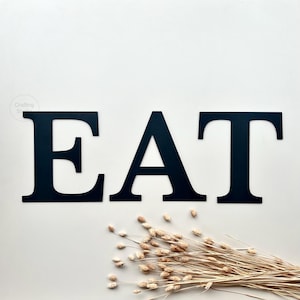Puede incluir: Letras metálicas negras que deletrean "EAT" sobre un fondo blanco. Las letras son grandes y en negrita, con una fuente moderna. Un manojo de espigas de trigo secas está dispuesto en la parte inferior. Ideal para la decoración de la cocina.
