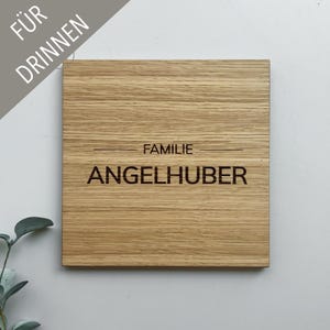 Puede incluir: Un letrero cuadrado de madera con las palabras "FAMILIE ANGELHUBER" grabadas en negro. El letrero tiene un patrón de vetas de madera natural y el texto "FÜR DRINNEN" está en la esquina superior izquierda.