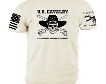 Puede incluir: Camiseta color crema con un gráfico negro que presenta una calavera con sombrero de vaquero, sables cruzados y el texto "U.S. Cavalry". La camiseta también muestra la bandera estadounidense en la manga izquierda y el texto "Because Even Marines Need Heroes."